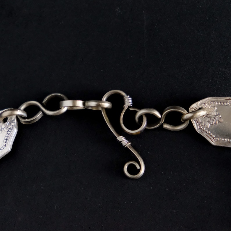 Puede incluir: Primer plano de un collar de cadena de plata con un cierre decorativo. La cadena est&aacute; hecha de mangos de cuchara unidos y el cierre es un bucle de alambre retorcido.