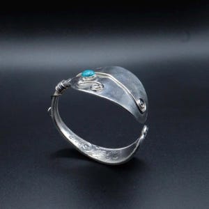Könnte beinhalten: Ein handgefertigtes silbernes Armband mit einem Türkisstein. Das Armband besteht aus recyceltem Besteck, mit einem Löffeldesign und dekorativen Wirbeln. Das Armband ist offen und verstellbar.