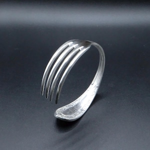 May include: A silver-toned metal bracelet fashioned from a bent fork. The tines of the fork curve upward, while the handle is curved into a circular shape to form the band. The handle features an embossed decorative pattern along its edges.