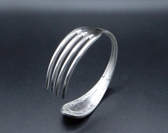 Puede incluir: Una pulsera de metal plateado hecha a partir de un tenedor doblado. Los dientes del tenedor se curvan hacia arriba, mientras que el mango está doblado en forma circular para formar la banda. El mango presenta un patrón decorativo en relieve.