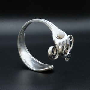 Può includere: Braccialetto d'argento realizzato con una forchetta riutilizzata con un accento di pietra opale bianca. I denti della forchetta sono arricciati in un design decorativo.
