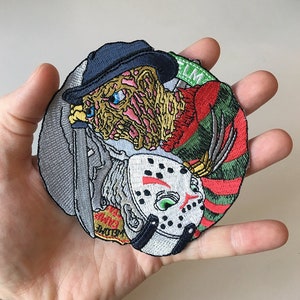 Puede incluir: Un parche bordado colorido con dos personajes de películas de terror, uno con una camisa a rayas rojas y verdes y una máscara blanca, y el otro con un sombrero negro y una máscara blanca con detalles rojos. El parche está sobre un fondo gris.