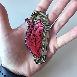 Peut inclure: Patch brodé représentant un cœur humain rouge à l'intérieur d'un cercueil en bois. Le cœur est détaillé avec des veines et des artères. Le cercueil est fermé et possède une bordure dorée.