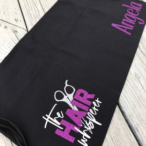 May include: Black apron with a purple glitter design that says "The Hair Whisperer" with silver scissors. The name "Angela" is written in purple glitter on the right side of the apron.