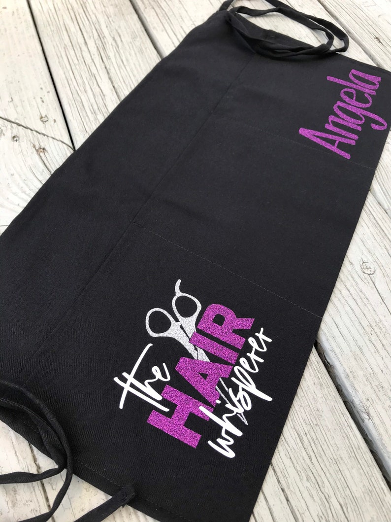 May include: Black apron with a purple glitter design that says "The Hair Whisperer" with silver scissors. The name "Angela" is written in purple glitter on the right side of the apron.