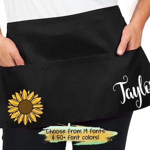 May include: Black apron with a sunflower design and the name "Taylor" in white script. The apron has a large pocket on the front. Choose from 14 fonts and 50+ font colors.