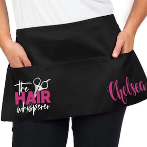 May include: Black hair stylist apron with a pink design that says "The Hair Whisperer" with a pair of scissors and the name "Chelsea" in pink.
