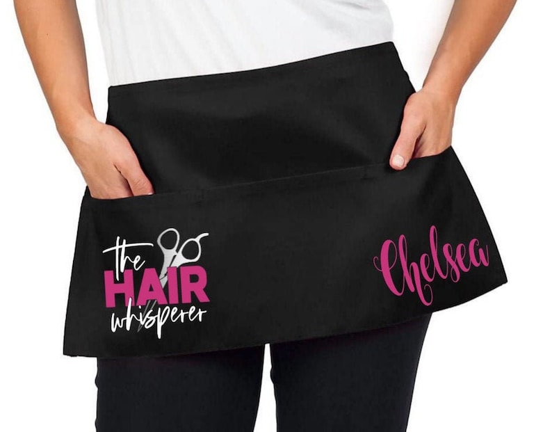 May include: Black hair stylist apron with a pink design that says "The Hair Whisperer" with a pair of scissors and the name "Chelsea" in pink.