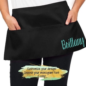 May include: Black waist apron with two front pockets. The apron has the name "Brittany" embroidered in a teal script font on the right side. The text "Customize your design! Choose your monogram font & color" is also visible.