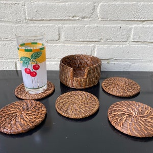 May include: A set of woven rattan coasters and a matching holder. A glass with a cherry design sits on one coaster. The coasters are round and have a natural brown color. The holder is cylindrical.