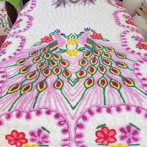 May include: A white crocheted bedspread with a pink and green peacock design. The peacock has yellow, red, and blue accents. The bedspread has a floral pattern around the edges.
