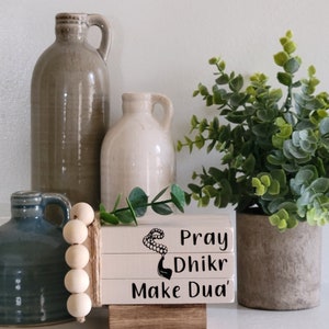 May include: A stack of three white wooden blocks with black text that reads "Pray Dhikr Make Dua'" and a black image of prayer beads. The blocks are sitting on a wooden base and are surrounded by a wooden bead garland, a gray ceramic pitcher, a white ceramic pitcher, and a potted green plant.