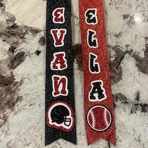 May include: Two black and red glitter ribbon banners with the names "Evan" and "Ella" in black letters. The Evan banner has a black football helmet and the Ella banner has a red baseball.