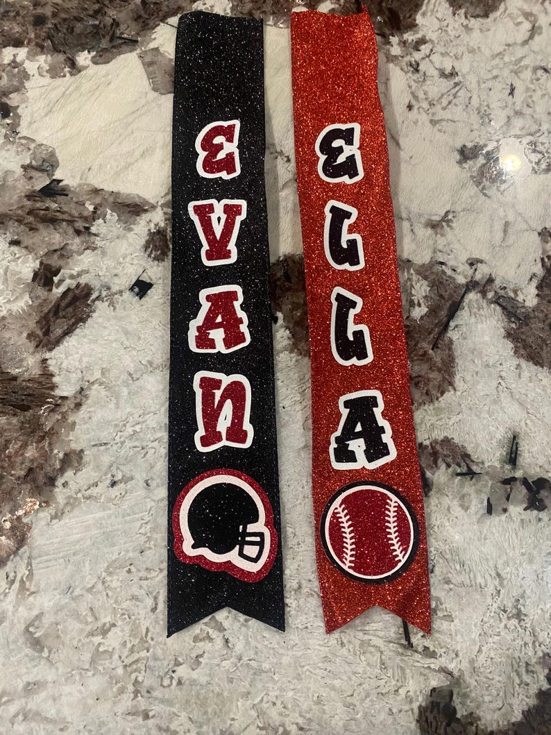 May include: Two black and red glitter ribbon banners with the names "Evan" and "Ella" in black letters. The Evan banner has a black football helmet and the Ella banner has a red baseball.