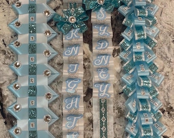 May include: Three blue and white ribbon sashes with the name "KYNLEY" spelled out in white letters. Each sash has a different design, one with a checkered pattern, one with a diamond pattern, and one with a bow pattern. The sashes are decorated with rhinestones and a football-shaped charm.