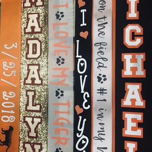 May include: Five orange and white fabric ribbons with text. The ribbons say "Madalyn", "I Love My Tiger", "I Love You", "#4 on the field #1 in my heart", and "Michael".