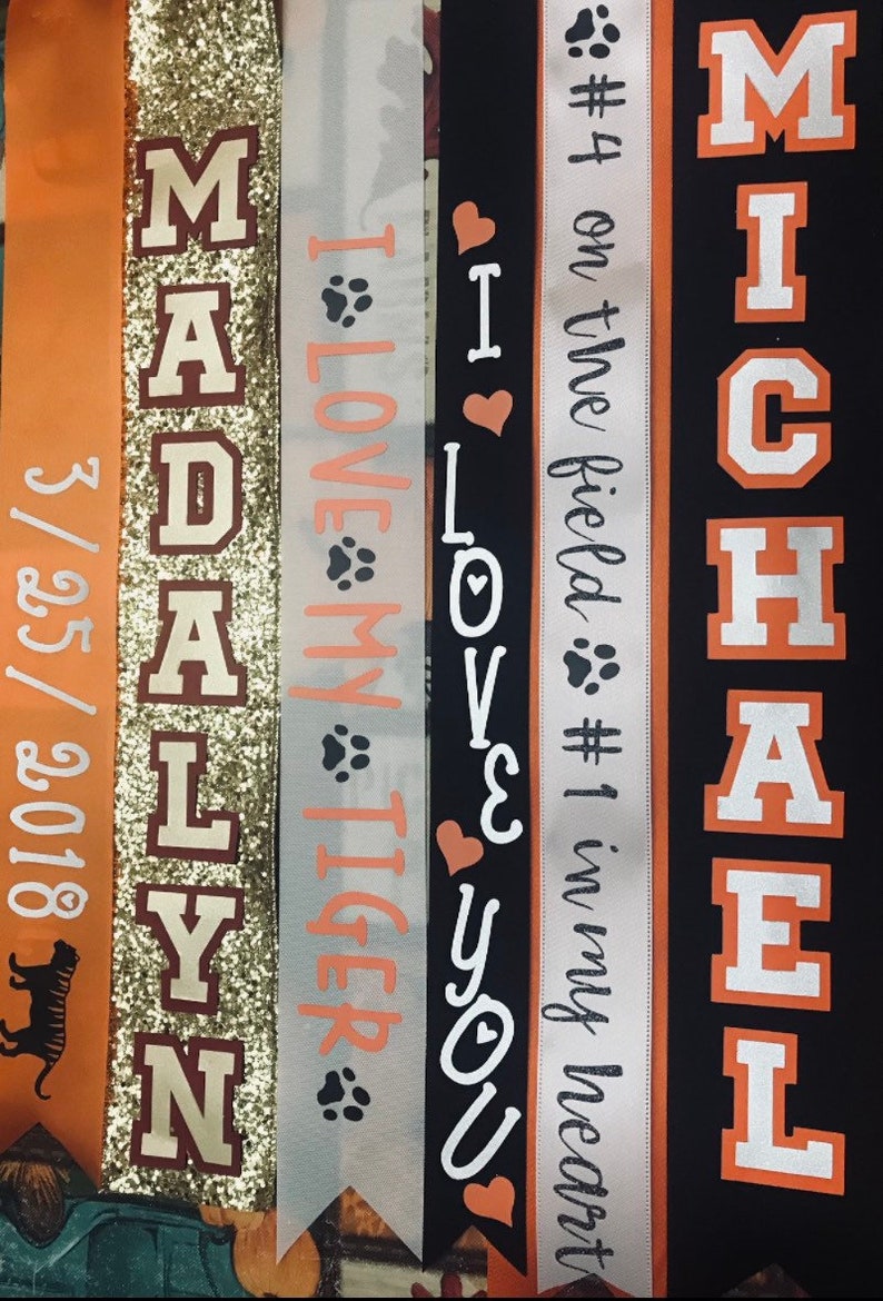 May include: Five orange and white fabric ribbons with text. The ribbons say "Madalyn", "I Love My Tiger", "I Love You", "#4 on the field #1 in my heart", and "Michael".