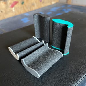 Könnte beinhalten: Eine Sammlung schwarzer Schaumstoff-Klettergriffe mit grauen und türkisfarbenen Akzenten. Die Griffe sind auf einer schwarzen Oberfläche angeordnet und zeigen ihr strukturiertes, ergonomisches Design. Im Hintergrund befindet sich eine Kletterwand.