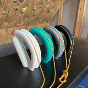 May include: A row of five climbing holds in white, teal, gray, and black, connected by a yellow rope. The holds have a curved, ergonomic design. The rope is knotted at the bottom. The holds are on a black surface.