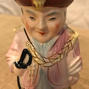 May include: A vintage ceramic pitcher shaped like a man wearing a yellow and red hat, a pink and blue jacket, and black shoes. He is holding a black object in his left hand.
