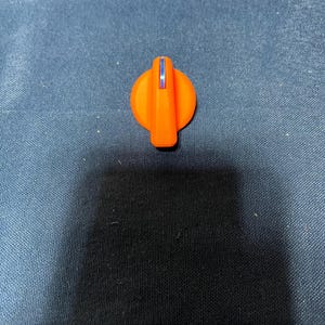 Puede incluir: Un mando de plástico naranja con un asa rectangular vertical y una franja azul. El mando es circular con un asa elevada. El objeto está sobre un fondo de tela azul oscuro.