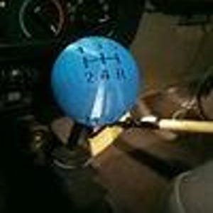 May include: A spherical blue gear shift knob featuring a black printed shift pattern with numbers 1, 2, 3, 4 and the letter R. The knob is mounted on a black metal shaft within a vehicle interior, positioned in front of a dark dashboard and gauge cluster.