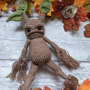 Pode incluir: Um brinquedo amigurumi de crochê marrom com folhas verdes para orelhas. O brinquedo está deitado em uma superfície de madeira branca com folhas de outono ao redor. O brinquedo tem uma expressão de raiva no rosto.