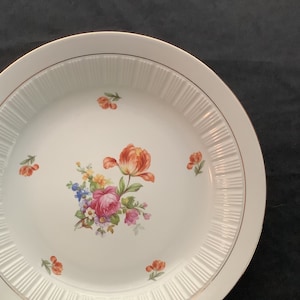 May include: White ceramic plate with a gold rim and a floral design featuring pink, red, and yellow flowers. The plate has a fluted edge.