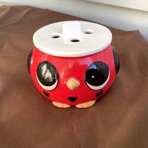 May include: A round, red ceramic container shaped like a bird with large black eyes, a small tan beak, and black heart-shaped accents. It features a white, circular lid with four holes, resembling a button, and small tan feet at the base.