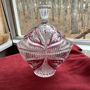 May include: A clear, cut-glass candy dish with a lid, featuring a geometric design with pink accents. The dish is diamond-shaped with a detailed, textured surface. The lid has a decorative handle. The dish sits on a red surface.