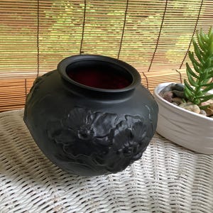 May include: A matte black decorative vase with a floral relief pattern. The interior is a deep red. Beside it is a white oval planter with a green succulent and small stones. The objects are on a white wicker surface.