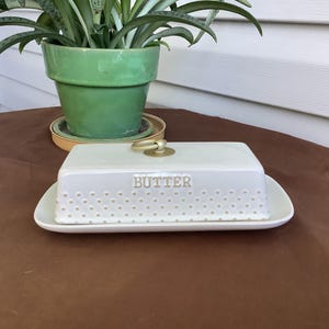 May include: A rectangular white ceramic butter dish with a matching lid featuring a gold-toned ring handle on top. The lid is embossed with the word BUTTER and a repeating pattern of small raised dots along the lower edge of the cover.