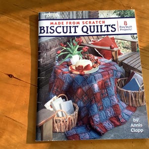May include: A book cover titled "Made From Scratch Biscuit Quilts" featuring a red, white, and blue quilt on a table. The cover also shows a basket, flowers, and a chair. The book has 8 rag quilt projects.
