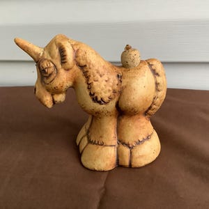 May include: A tan ceramic unicorn figurine with a speckled texture and dark brown etched detailing. The unicorn features a single horn, a rounded body, and a small spherical accent on its back, standing on four thick, block-like legs.