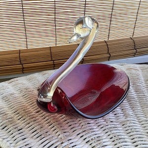 May include: A decorative glass swan figurine with a deep red body and a clear, curved neck and head. The swan is resting on a white woven surface, with a bamboo blind in the background.