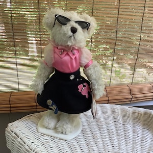 May include: A white teddy bear wearing black cat-eye sunglasses, a pink top with rhinestone accents, and a black skirt with a pink poodle applique. The bear is standing on a white base.