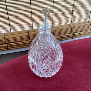 May include: A clear, cut-glass oil lamp with a decorative, diamond-patterned design. The lamp has a long, white wick extending from the top. The lamp is sitting on a red surface.