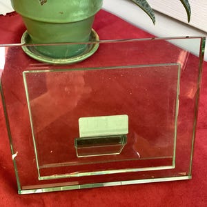 May include: A rectangular clear glass picture frame featuring a wide border and a central opening for a photograph. The frame includes a small, rectangular, light-colored adhesive label attached to the center of the glass surface.