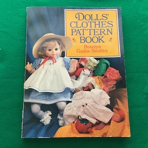 May include: A book titled "Dolls' Clothes Pattern Book" by Roselyn Gadia-Smitley. The cover features a doll wearing a blue and white gingham dress with a white apron and a straw hat. The doll is surrounded by fabric scraps and sewing supplies.