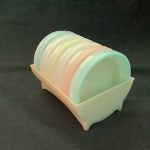 May include: A set of six pastel-colored coasters in a white plastic holder. The coasters are stacked in a rainbow pattern, with the top coaster being a light blue color.