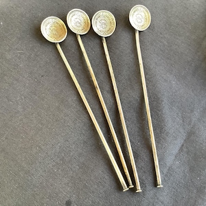 May include: Four antique silver cocktail stirrers with round, coin-like tops featuring embossed designs. The stirrers have long, slender stems and are arranged diagonally against a dark gray background. The tops have a detailed pattern.