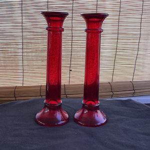 May include: Two tall, red glass candle holders. Each has a flared base, a long, cylindrical body, and a wide, slightly flared top. The candle holders are displayed on a dark surface against a backdrop of a bamboo blind.