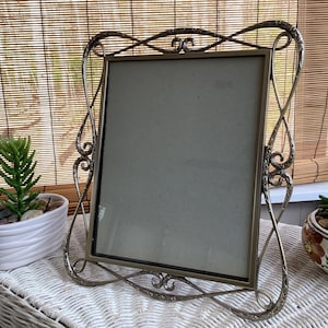 May include: An ornate, antique-style metal picture frame with a rectangular opening. The frame has a decorative, swirling design and a metallic finish. The frame is displayed on a white wicker surface, with a small succulent plant in a white pot to the left.