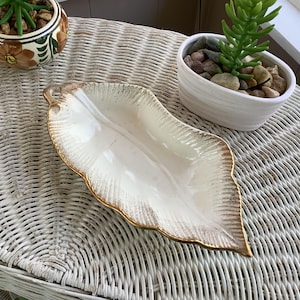 May include: A white, leaf-shaped ceramic dish with a gold-colored trim. The dish is on a white wicker table, with two small potted plants in the background. The dish has a textured surface.