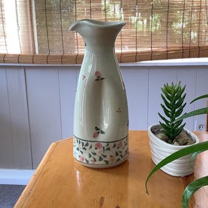 May include: A tall, cream-colored ceramic pitcher with a flared spout and a narrow neck. The body features a painted pattern of small pink fruits and green leaves, with a decorative band of similar foliage encircling the base of the vessel.