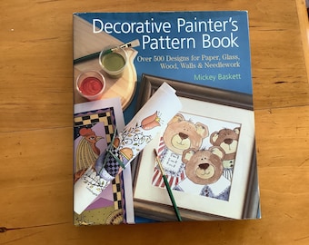 May include: A book titled "Decorative Painter's Pattern Book" with a blue cover and the text "Over 500 Designs for Paper, Glass, Wood, Walls & Needlework" and "Mickey Baskett". The book cover features an illustration of three bears and a paintbrush.