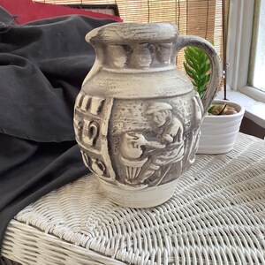 May include: A vintage ceramic pitcher with a handle, featuring a relief design of a craftsman at work. The pitcher is primarily off-white with gray accents, and stands on a white wicker table. A small succulent plant is visible.