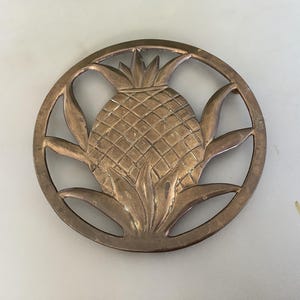 May include: A circular brass trivet featuring an openwork design of a pineapple in the center. The metal has a warm, aged patina with visible surface texture and a cross-hatched pattern on the fruit body, surrounded by stylized leaves.