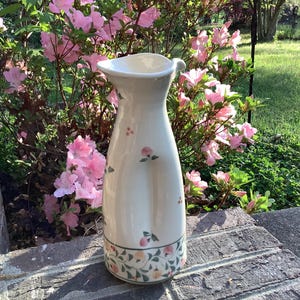 May include: A tall, off-white ceramic pitcher with a flared top and a small handle. The pitcher is decorated with small pink floral accents and a band of floral and leaf patterns at the base. The background features pink azalea flowers.