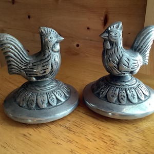May include: Two silver-toned metal rooster figurines, each featuring a detailed bird perched atop a circular base decorated with a leaf-like pattern. The roosters have textured feathers and distinct combs, with a metallic finish and dark accents.